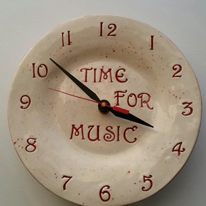 May include: A white ceramic clock with red numbers and a black clock hand. The clock face has a red splatter pattern and says "TIME FOR MUSIC".