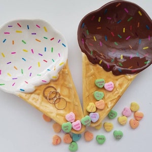 May include: Two ceramic ice cream cone dishes, one with white frosting and rainbow sprinkles, the other with brown chocolate frosting and sprinkles. Both cones are filled with colorful candy hearts.