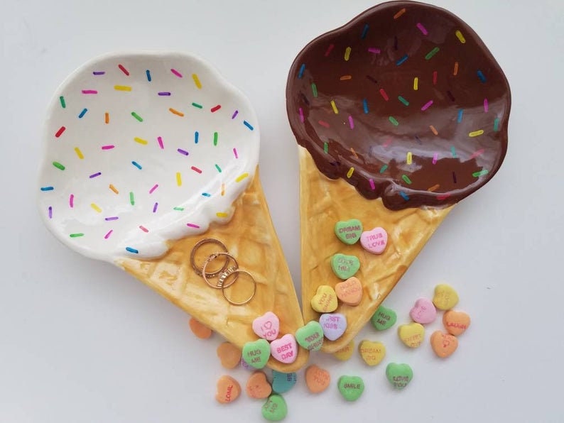 May include: Two ceramic ice cream cone dishes, one with white frosting and rainbow sprinkles, the other with brown chocolate frosting and sprinkles. Both cones are filled with colorful candy hearts.