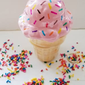 May include: A pink ceramic ice cream cone with colorful sprinkles. The cone is sitting on a white surface with more sprinkles scattered around it.