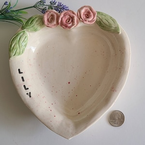 May include: A white heart-shaped ceramic dish with a pink and green floral design. The dish has a speckled glaze and the word "Love" written on the side.