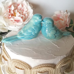 May include: Two blue ceramic birds perched on a white cake with a cream-colored, rope-like trim. The birds are facing each other and appear to be kissing.