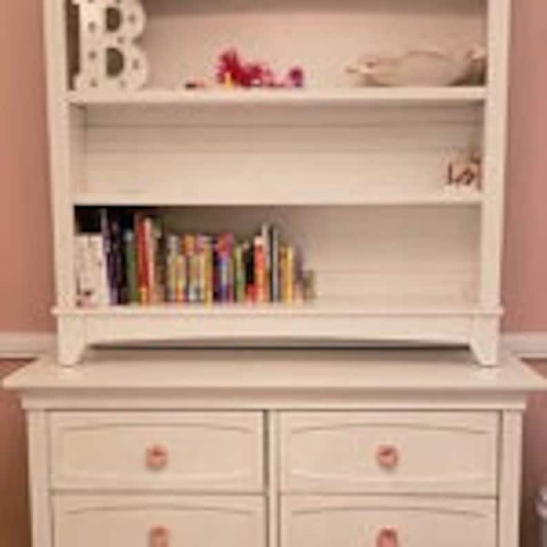 May include: White wooden bookcase with four shelves, the top shelf has a decorative letter B, the second shelf is empty, the third shelf has a collection of books, and the bottom shelf is part of a white dresser with four drawers and pink knobs.