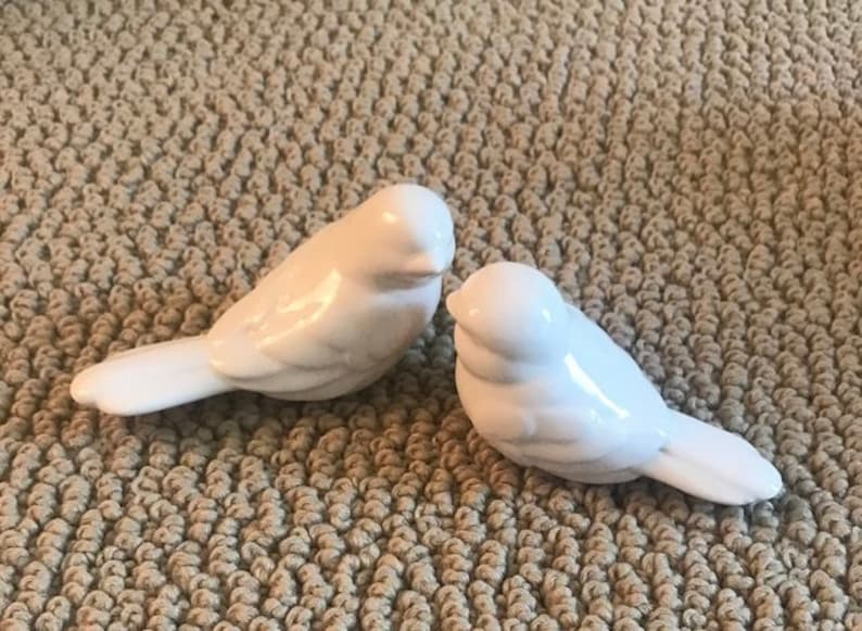 May include: Two small, white ceramic bird figurines with smooth, glossy finishes. Each bird is depicted in a resting position with wings tucked against its body, a rounded head, and a short, pointed beak, showcasing a clean and simple design.