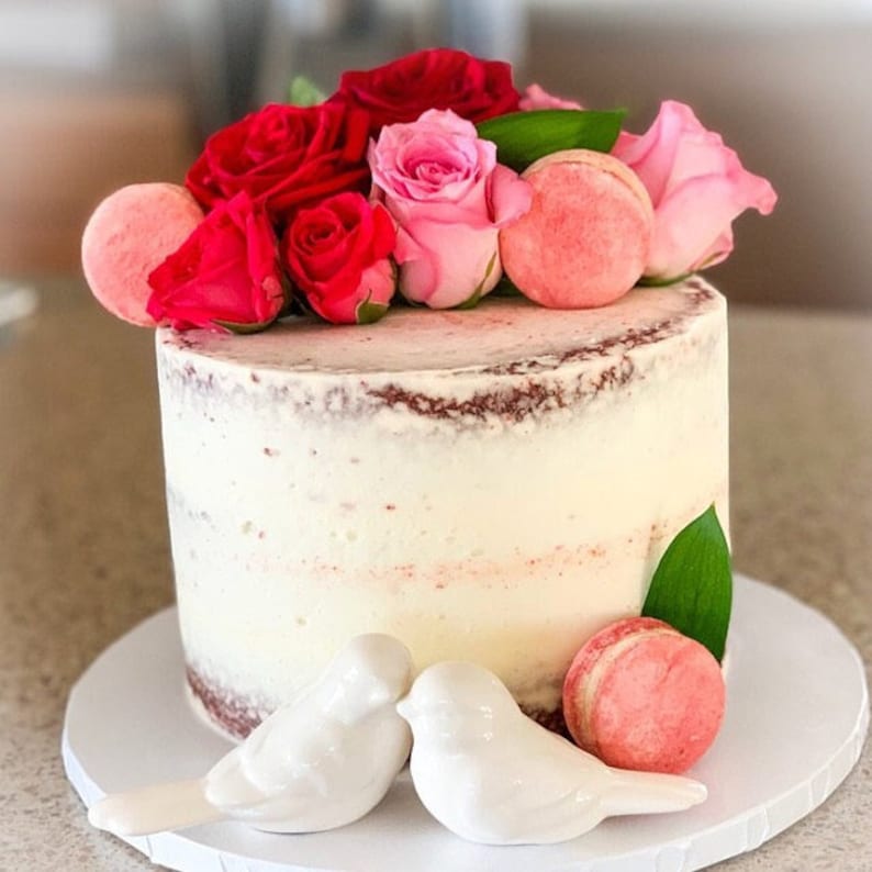 May include: A round, semi-naked cake with white frosting, topped with vibrant red and soft pink roses, green leaves, and round pink macarons. Two small, white ceramic bird figurines are placed at the base, alongside an additional pink macaron.