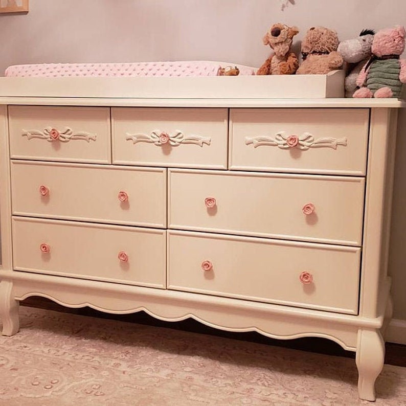 May include: A white dresser with six drawers and a changing table top. The dresser has a scalloped base and ornate floral-shaped drawer pulls.