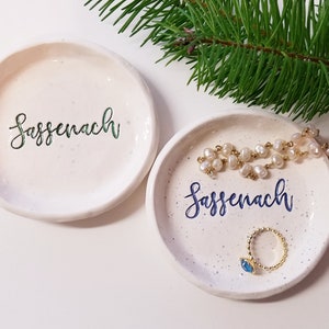 May include: Two white ceramic trinket dishes with a speckled finish. Both dishes have the word "Sassenach" written in cursive on them. One dish has the word in green and the other in blue. The dish with the blue writing has a gold ring with a blue stone and a pearl necklace on it.