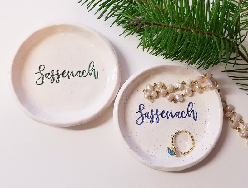May include: Two white ceramic trinket dishes with a speckled finish. Both dishes have the word "Sassenach" written in cursive on them. One dish has the word in green and the other in blue. The dish with the blue writing has a gold ring with a blue stone and a pearl necklace on it.