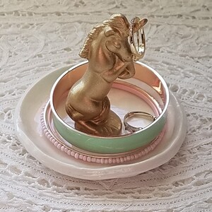May include: A gold-colored unicorn ring holder with a white ceramic dish base. The unicorn holds two rings, and several bracelets in shades of pink and mint green are stacked on the dish.