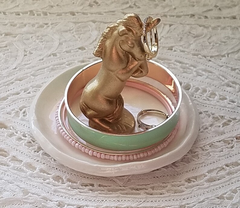 May include: A gold-colored unicorn ring holder with a white ceramic dish base. The unicorn holds two rings, and several bracelets in shades of pink and mint green are stacked on the dish.