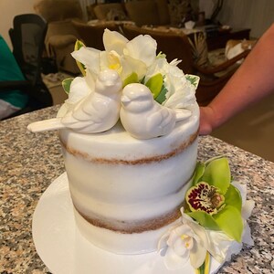May include: A semi-naked cake with white frosting and visible cake layers, topped with two white ceramic bird figurines and white flowers. A cluster of green and white orchids is attached to the base of the cake on a white circular cake board.
