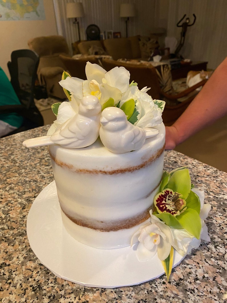 May include: A semi-naked cake with white frosting and visible cake layers, topped with two white ceramic bird figurines and white flowers. A cluster of green and white orchids is attached to the base of the cake on a white circular cake board.
