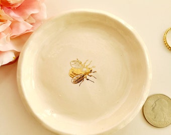 Puede incluir: Un pequeño plato de cerámica blanca con un diseño de abeja dorada. El plato es aproximadamente del tamaño de una moneda de euro.