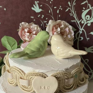 May include: A decorative cake topper featuring two ceramic birds, one green and one cream-colored, perched atop a white cake. The cake is adorned with a heart-shaped embellishment and floral accents, perfect for a wedding or anniversary celebration.