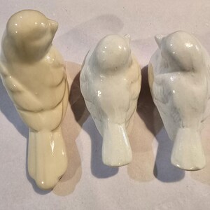 May include: Three small ceramic bird figurines arranged in a row. The bird on the left is a creamy off-white color, while the two birds on the right are a bright, glossy white. Each bird is depicted from a top-down view, showing wings and tails.