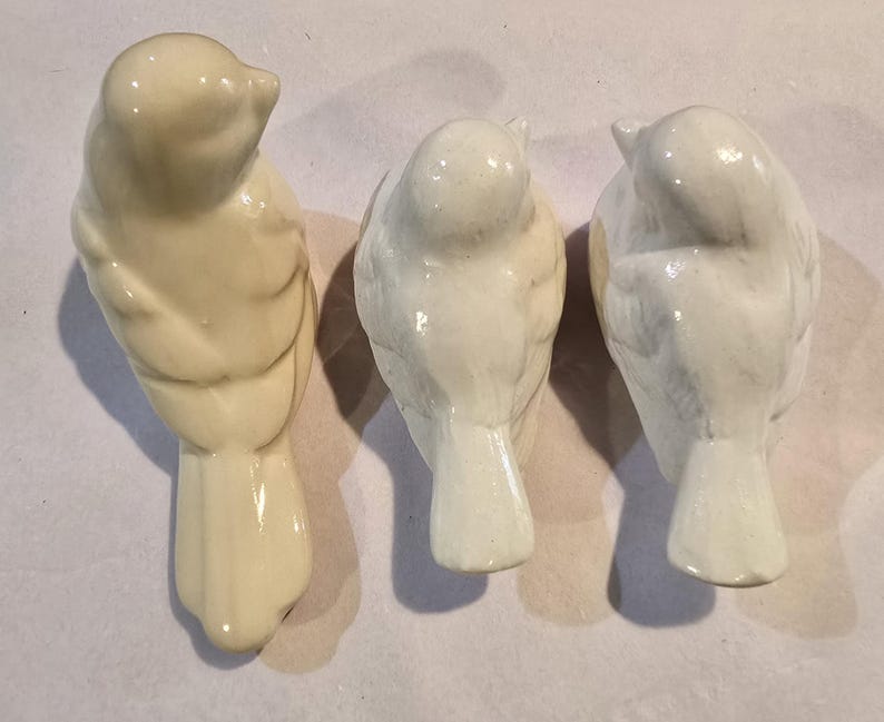 May include: Three small ceramic bird figurines arranged in a row. The bird on the left is a creamy off-white color, while the two birds on the right are a bright, glossy white. Each bird is depicted from a top-down view, showing wings and tails.