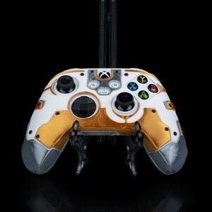 May include: A white and gold Xbox controller with a custom design. The controller is on a black stand.