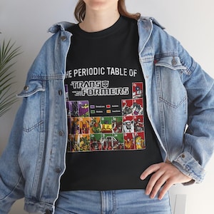 May include: Black t-shirt featuring "The Periodic Table of Transformers" design. The graphic includes various Transformers characters arranged in a periodic table format, with Autobots and Decepticons color-coded. The shirt is worn with a blue denim jacket.