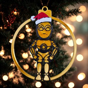 May include: A gold and black Christmas ornament featuring a robot character wearing a Santa hat. The ornament is in the shape of a circle and is hanging on a Christmas tree. The robot has a gold body and black accents.