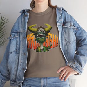 Peut inclure: T-shirt marron avec un monstre de dessin animé aux cornes jaunes, au corps vert et aux dents pointues. Le monstre est devant le texte orange "RRRAAAGGH!". Le t-shirt est porté avec une veste en jean bleue.