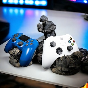 Pode incluir: Um controle PlayStation azul e um controle Xbox branco repousam sobre bases rochosas detalhadas. Uma pequena estatueta preta de um soldado apontando uma arma é posicionada entre os controles. Os controles estão em uma superfície branca com um fundo desfocado.