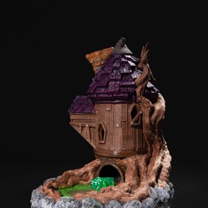 May include: A 3D printed tabletop game dice tower shaped like a small house with a purple roof and a tree trunk base. The tower has a small opening at the bottom where dice can be rolled out. A green die is visible inside the tower.