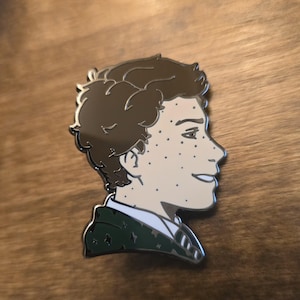 May include: A metal pin featuring a side profile of a person with brown hair and freckles. The pin has a silver outline and a green and white jacket. The pin is on a wooden surface.