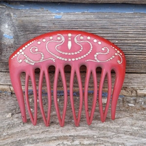 Puede incluir: Un peine de plástico rojo con un diseño decorativo en plata. El peine tiene una parte superior curva y siete dientes.