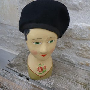 May include: A solid black beret with a smooth texture and a matching black fabric trim along the base. The beret is displayed on a painted mannequin head featuring green eyes, red lips, and a small floral design on the neck area.
