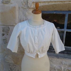 May include: A white cropped blouse featuring three-quarter length sleeves, a round neckline with delicate lace trim, and a front button-down closure. The garment is made of a lightweight fabric with gathered detailing at the waist and neckline.