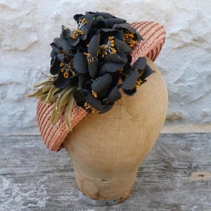 May include: A peach-colored woven headband featuring a large cluster of dark charcoal-colored fabric flowers with bright yellow centers. The floral arrangement is positioned on the side of the headband, which is displayed on a beige mannequin head.