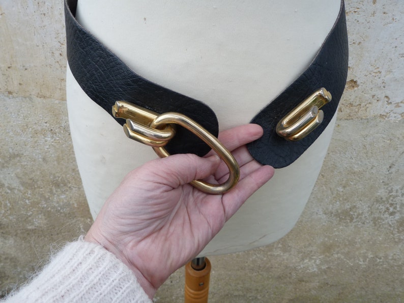 May include: A black leather belt with a gold metal buckle. The buckle is a large, intricate design with multiple loops and curves.