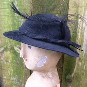 May include: A black felt hat with a wide brim and a feather accent. The hat is sitting on a wooden mannequin head.