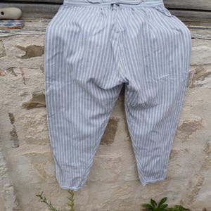 Op de afbeelding: Lichtblauwe en witte broek met verticale strepen, voorzien van een hoge taille en een verstelbare band aan de achterzijde. De tailleband heeft vier kleine witte knopen en een centrale verstelbare lip, met toelopende pijpen.