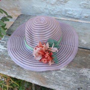 May include: A light purple sun hat with a wide brim and a decorative floral arrangement. The hat features a striped design and a light green ribbon. The flowers are in shades of orange and pink. The hat is resting on a weathered wooden surface.