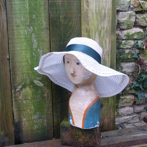 May include: A white and green painted wooden mannequin head wearing a white straw hat with a green ribbon. The hat has a wide brim and is decorated with white stitching.