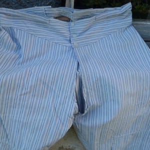 Op de afbeelding: Lichtblauwe en witte gestreepte vintage broek. De broek heeft een hoge taille, een knoopsluiting en een split aan de voorkant. De stof lijkt van lichtgewicht katoen of linnen te zijn. Het kledingstuk is waarschijnlijk antiek of vintage.