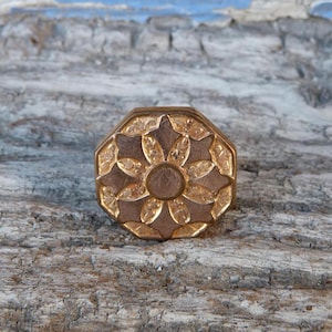 May include: A gold-toned octagonal metal knob with a decorative floral design. The knob has a raised center and a textured surface.