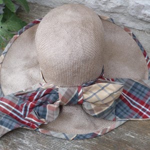 May include: A beige sun hat with a wide brim and a matching crown. The brim is trimmed with a plaid fabric in red, blue, and tan. A large plaid bow adorns the front of the hat, adding a decorative touch.