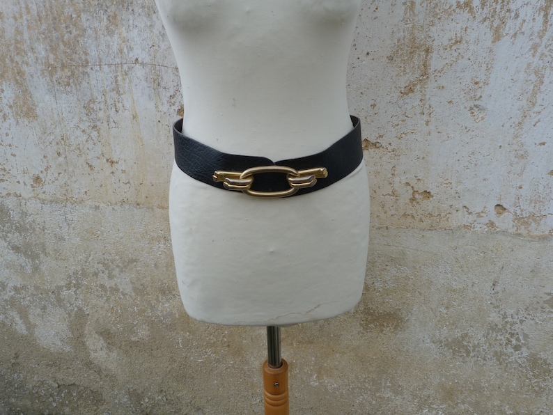 May include: A black leather belt with a gold-toned buckle featuring a large oval link design.