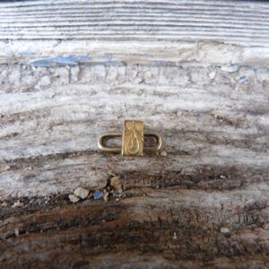 Puede incluir: Un pequeño cierre dorado con un diseño grabado y ornamentado. El cierre tiene una pieza central rectangular con un patrón curvo en forma de lágrima y dos bucles redondeados a cada lado. El fondo es una superficie de madera envejecida.