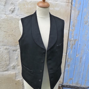 Könnte beinhalten: Eine schwarze Weste mit Schalkragen und Knopfverschluss, präsentiert auf einer Schaufensterpuppe. Die Weste hat zwei Vordertaschen und eine taillierte Passform. Der Hintergrund zeigt eine Steinmauer und eine blaue Holztür.