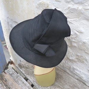 May include: A black hat with a textured, woven appearance and a wide, slightly asymmetrical brim. The crown features a structured, folded design with a small, decorative fabric bow detail positioned on the side of the brim near the base.