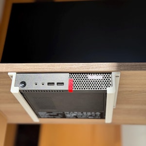 Könnte beinhalten: Ein schwarzer Computer-Tower mit weißem Rand ist unter einem Holztisch montiert. Der Computer hat ein ThinkCentre-Logo auf der Vorderseite und mehrere Anschlüsse an der Seite.