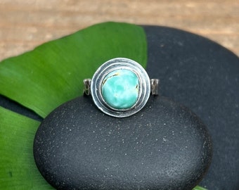 May include: A silver ring with a turquoise gemstone set in a circular design. The ring is resting on two black stones.