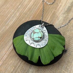 May include: A silver necklace with a turquoise stone pendant set in a silver filigree design. The pendant is shaped like a teardrop and is surrounded by a circle of smaller silver circles. The necklace is lying on a green leaf.