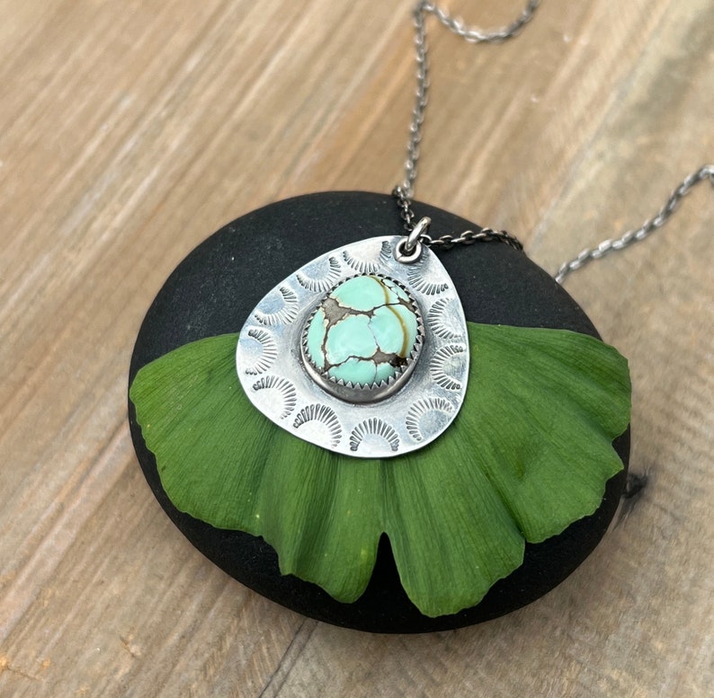 May include: A silver necklace with a turquoise stone pendant set in a silver filigree design. The pendant is shaped like a teardrop and is surrounded by a circle of smaller silver circles. The necklace is lying on a green leaf.