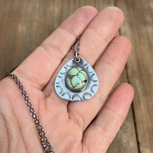 May include: A silver necklace with a teardrop-shaped pendant featuring a green turquoise stone surrounded by a decorative silver border. The pendant has a scalloped edge.