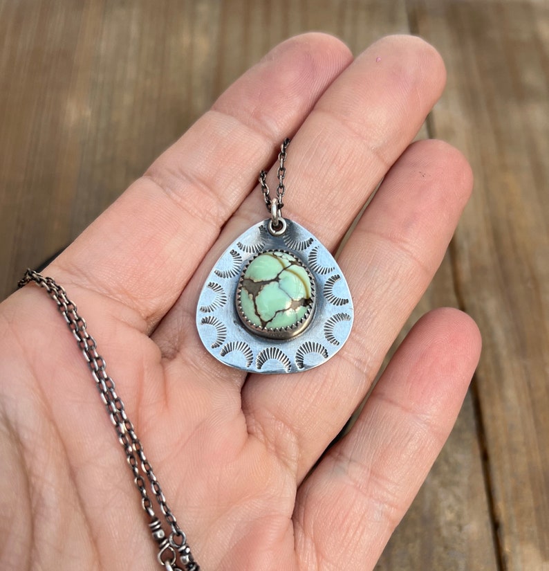 May include: A silver necklace with a teardrop-shaped pendant featuring a green turquoise stone surrounded by a decorative silver border. The pendant has a scalloped edge.