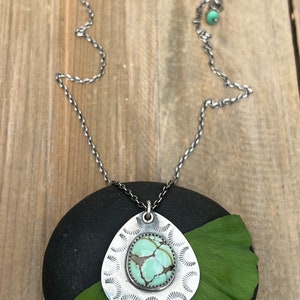 May include: A silver necklace with a teardrop-shaped pendant featuring a green turquoise stone set in a silver frame. The pendant is surrounded by a decorative silver design. The necklace is on a black stone with a green leaf.